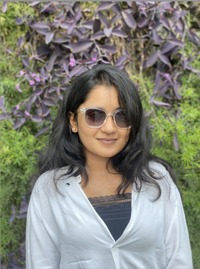 Krishanee Prajapati posing in front of a lush green and purple foliage wall, wearing sunglasses and a white shirt with a black lace top underneath.